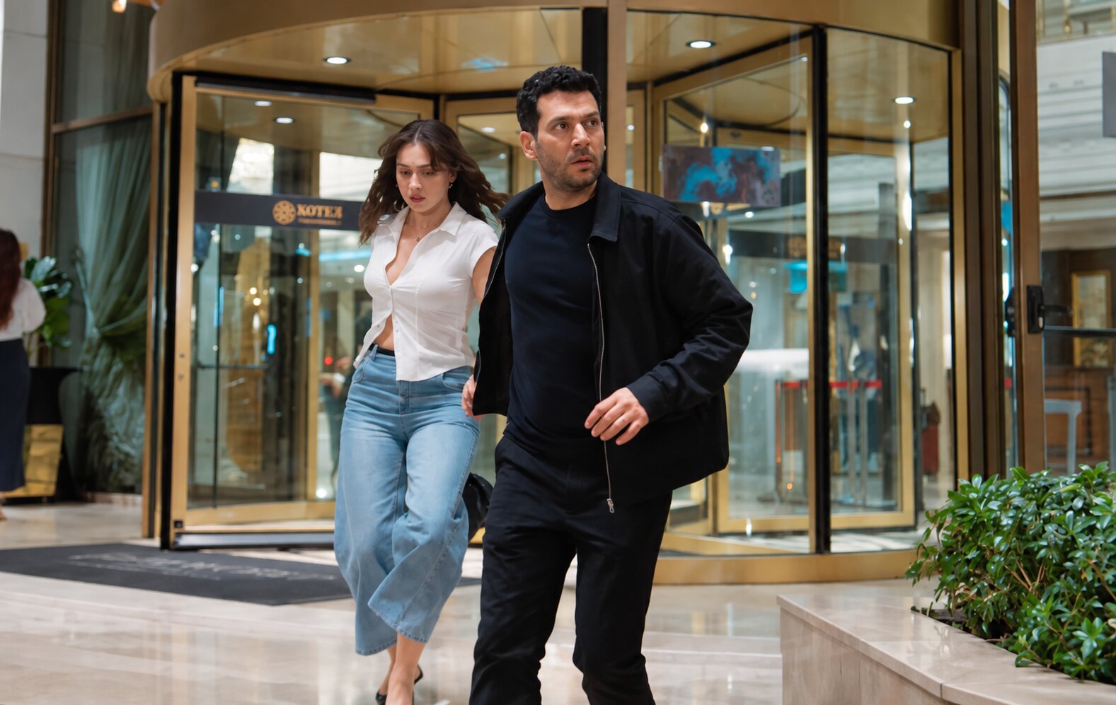Murat Yıldırım as Serhat Tecer holding Cemre Baysel as Zeynep Candan while running anxiously through a hotel entrance in Sins and Roses Season 2 (Güller ve Günahlar).